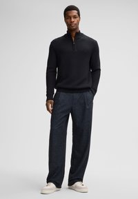 Zwarte gebreide trui met halve rits kraag, gecombineerd met losse, donkergepatroneerde broek en beige sneakers. Focus op textuur en kleurcontrast.