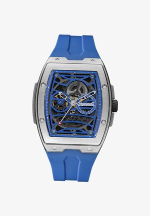 Montre automatique carrée argentée et bleue avec engrenages exposés, couronne noire, aiguilles blanches, trotteuse à pointe rouge et bracelet en silicone bleu.