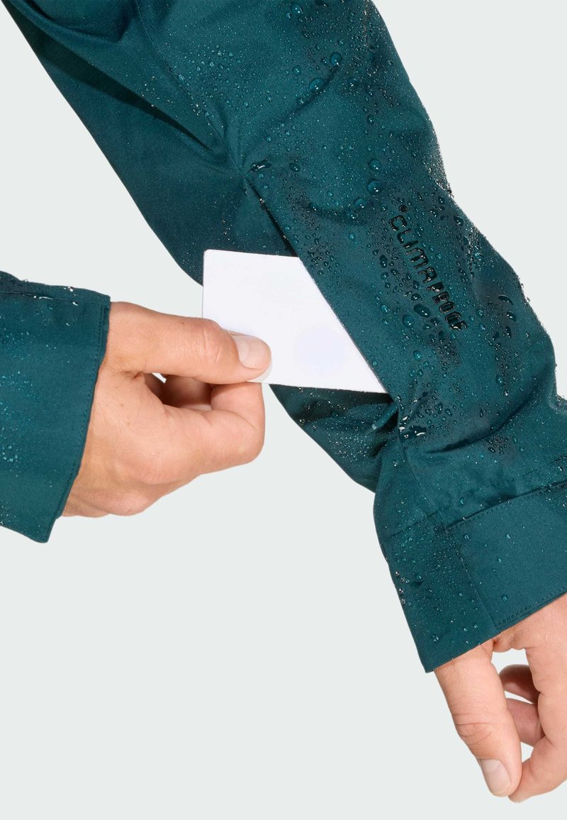 Türkisfarbene Jacke mit Wassertröpfchen an einem Arm, Hand legt eine weiße Karte in eine Brusttasche. Das Material scheint wasserdicht zu sein und hat eine glatte Textur.