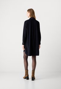 Donna con capelli di lunghezza spalla, che indossa un abito a camicia lungo con maniche nere, collant neri trasparenti e stivaletti alla caviglia in suede marrone, in piedi con la schiena rivolta.