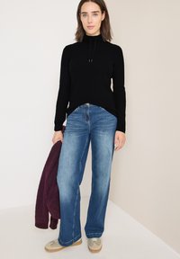 Maglione nero a coste con colletto alto, jeans blu a gamba larga e scarpe grigie; tiene in mano una giacca bordeaux. Design minimalista e stile casual.