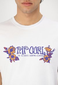 White cotton T-shirt with crew neckline, featuring colorful floral graphics in orange and purple, and the text "RIP CURL THE ULTIMATE SURFING COMPANY."