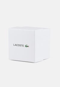 Vit rektangulär låda med Lacoste-logotypen och en alligatordiagram i grönt och rött, med en slät, matt yta.