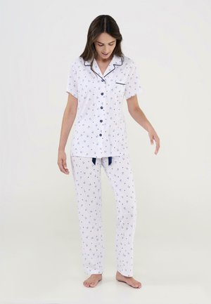Mujer con pijama blanco de manga corta con botones y pantalones largos a juego con pequeños estampados florales azules, de pie descalza.