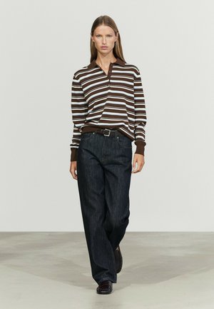 Donna che cammina con una maglia a righe marroni e bianche a maniche lunghe, jeans scuri a gamba larga, cintura nera e scarpe scure su sfondo neutro.
