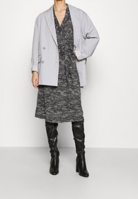 Grå överdimensionerad blazer över en klänning med svart och vitt mönster, kombinerad med knähöga svarta klackstövlar, vilket skapar en lager på lager-look.