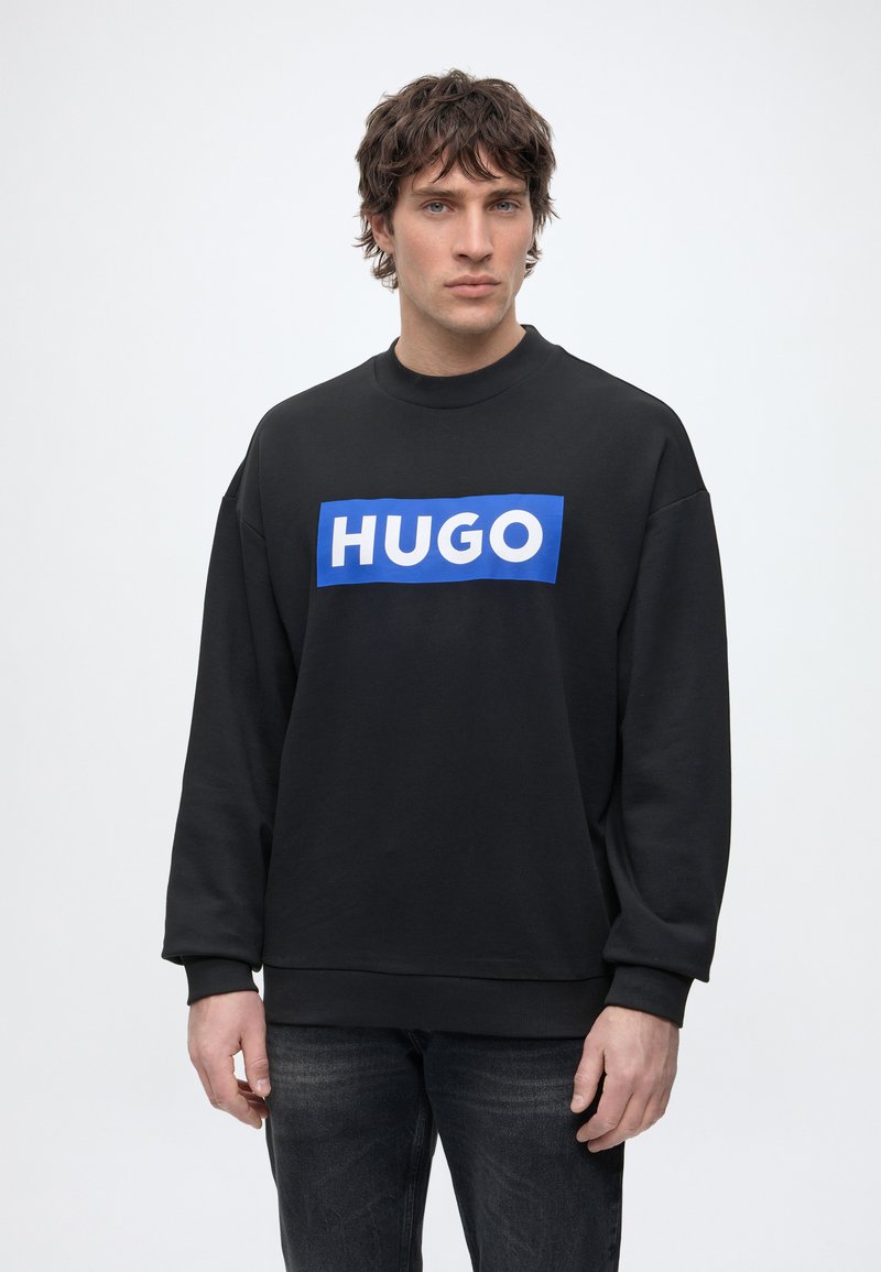 Mann mit dunklen lockigen Haaren, der einen schwarzen Pullover mit blauem "HUGO"-Logo und dunkle Jeans trägt, steht vor einem schlichten hellen Hintergrund.