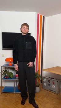 Νεαρός άνδρας με τατουάζ, ντυμένος με μαύρα ρούχα, στέκεται σε ένα δωμάτιο με τηλεόραση, πορτοκαλί λαμπατέρ, φυτά, ένα μπάσκετ και μια ασημένια βαλίτσα καλυμμένη με αυτοκόλλητα.