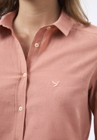 Camicia color salmone con colletto, dal texture morbida, caratterizzata da bottoni decorativi e un piccolo colibrì ricamato vicino al petto.