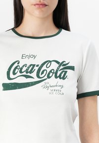 White t-shirt with green accents and a vintage Coca-Cola logo. Text reads "Enjoy Coca-Cola So Refreshing Served Ice Cold."