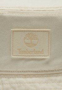 Beige stof med vandret ribbet tekstur, der har et debosseret Timberland-logo og trædesign på en rektangulær lap.