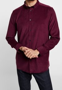 Bordeauxfarbenes Cordhemd mit langen Ärmeln, Button-Down-Kragen und schwarzen Knöpfen. Der Stoff hat eine weiche Textur mit dezenten vertikalen Linien.