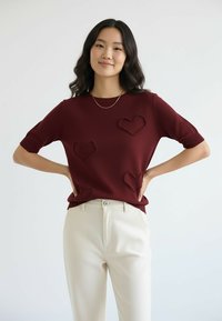 Bordowy sweter dziany z krótkimi rękawami, ozdobiony trzema fakturowanymi aplikacjami w kształcie serc; zestawiony z białymi spodniami i delikatnym złotym naszyjnikiem.