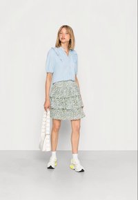 Camisa azul-claro com colarinho frisado, combinada com uma saia verde floral em camadas, sapatilhas brancas e uma bolsa tote bege.