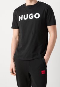 Camiseta de algodón negra con un gran logotipo en blanco "HUGO" en la parte frontal. Diseño de cuello redondo con mangas cortas, textura suave y corte relajado.