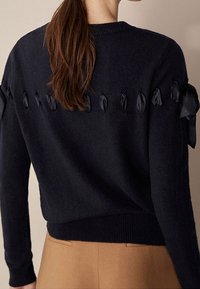 Tummansininen neulottu neulepaita, jossa on teksturoitu kangas, satiininauha yksityiskohdat olkapäillä ja resoroitu helma.