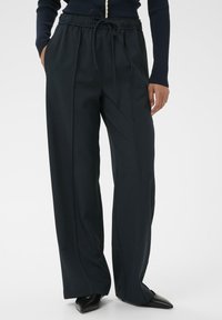 Vrouw in een zwarte wijde broek met plooien aan de voorkant, touwtje in de taille, zwarte puntige schoenen en een geribbelde lange mouwtop.