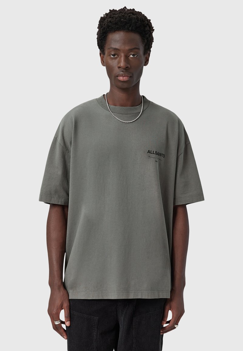 Camiseta de gran tamaño en gris claro con cuello redondo, mangas cortas y pequeño logo negro "ALLSAINTS" en el pecho. Textura de algodón suave.