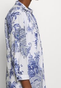 Uomo che indossa una camicia bianca con un intricato motivo paisley blu e un stampa architettonica, mostrato di lato su uno sfondo neutro.