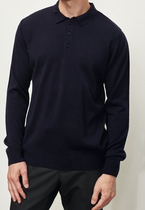 Uomo che indossa una polo a maniche lunghe color blu navy scuro con tre bottoni e pantaloni scuri, su sfondo chiaro e uniforme.