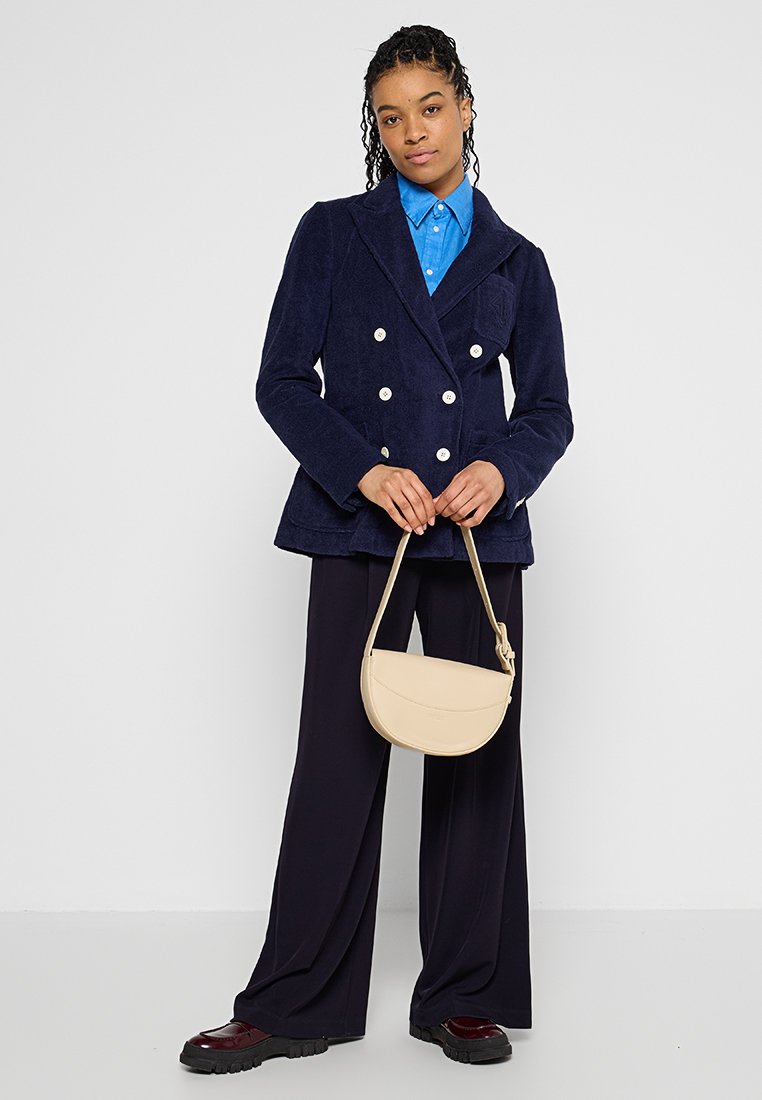 Granatowa kurtka z kordurowego materiału, zapinana na podwójne guziki, z dużymi białymi guzikami, niebieska koszula pod spodem oraz beżowa torba na ramię, w połączeniu z ciemnymi spodniami.