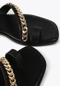Mustat sandaalit, joissa on litteä pohja, kaksi leveää remmiä ja kultaketjuakseli yläosassa. Sujuva pinta ja tyylikäs muotoilu.