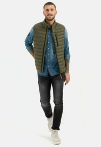 Olivgrüner Steppweste über einem Jeanshemd, kombiniert mit dunklen Jeans und hellen Sneakers. Die Weste hat eine Reißverschlusstasche und ein gestepptes Design.