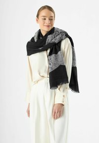 Écharpe noire et grise à motifs drapée sur les épaules, avec des bords à franges et une texture douce. Portée avec une blouse blanche et un pantalon.