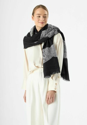 Schwarz-weiß gemusterter Schal, der über den Schultern drapiert ist, mit Fransen und einer weichen Textur. Getragen mit einer weißen Bluse und Hose.