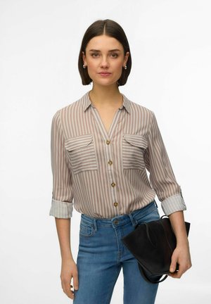 Femme aux cheveux courts et foncés portant une chemise boutonnée beige à rayures et un jean bleu, tenant un sac à main noir sur fond blanc.