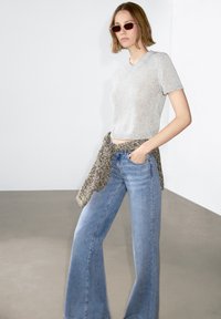 Maglione grigio a maniche corte in maglia, abbinato a jeans blu a gamba larga e una sciarpa testurizzata legata in vita. Superficie liscia, vestibilità rilassata.