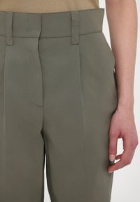 Khaki trousers featuring a flat waistband, two front pleats, and subtle stitching details. Smooth fabric with a slight sheen.