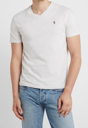 Homme portant un t-shirt gris clair à col en V avec un petit logo de joueur de polo sur la moitié gauche de la poitrine et un jean bleu, debout devant un fond uni.