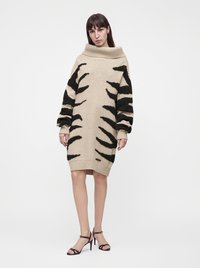 Femme portant une robe pull en maille beige et noire à motifs, manches longues, et des sandales noires à talons hauts, debout devant un fond uni.