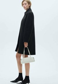 Čierny oversized kabát s golierom, doplnený bielou kabelkou. Modelka má na sebe čierne ponožky a lodičky, predvádzajúc moderný siluetu.