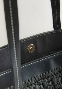 Sac à main noir en tissu tissé avec une texture tissée, une garniture en cuir noir avec des coutures blanches et un fermoir à snap en métal doré.