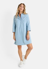 Robe en denim bleu clair avec broderies florales, col et manches trois-quarts. Le mannequin porte des baskets blanches, mettant en avant une coupe décontractée.