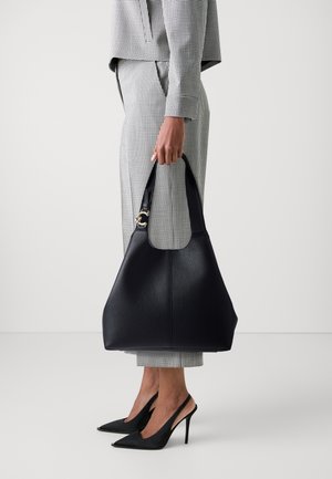 Dark navy leather handbag with textured surface and wide handle, held by a person wearing a checkered suit and black pointed heels.