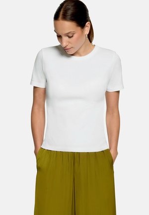 Femme aux cheveux bruns en queue de cheval portant un haut blanc uni à manches courtes et un pantalon taille haute vert olive, les mains dans les poches.