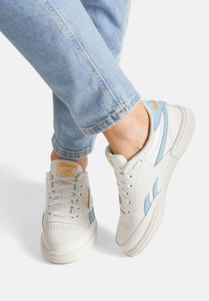 Balti ādas sporta apavi ar gaiši zilām detaļām, perforācijām, teksturētu zolīti un oranžu zīmolu mēlē un papēdī.