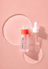 Klar glass serumflaske med gradient oransje topp, pipettehette og hvit etikett. Omgitt av dråper på en myk rosa bakgrunn.