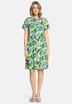 Vrouw draagt een knielange jurk met korte mouwen, bedrukt met een groenblauw tropisch bladpatroon, en witte sandalen met zwarte studs.