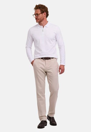 Mann mit Brille, der ein weißes langärmliges Poloshirt, beige Hose, braunen Gürtel und dunkelbraune Schuhe trägt, steht vor einem schlichten Hintergrund.