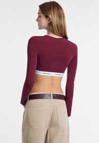 Bordeaux lange mouwen crop top met een ronde halslijn, waardoor een witte elastische band zichtbaar is met het logo "GUESS JEANS". Gecombineerd met lichtbeige hoge taillebroeken.