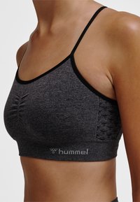 Tułów kobiecy w ciemnoszarym, bezszwowym sportowym biustonoszu z cienkimi czarnymi ramiączkami i logo „hummel” na opasce, widziany z boku.