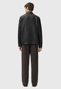 Zwarte leren jack met een gladde afwerking, grote kraag en een ontspannen pasvorm, gecombineerd met losse donkere broeken. Achteraanzicht.