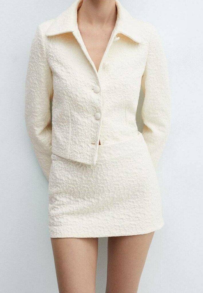 Femme portant une tenue deux pièces texturée crème avec une veste boutonnée à col et une mini-jupe taille haute sur un fond uni.
