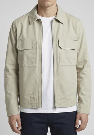 Man met een beige jas met rits en twee klepzakken op de borst, gedragen over een wit T-shirt en donkere broek, staand tegen een grijze achtergrond.