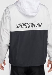 Czarna i biała kurtka sportowa z kapturem, z napisem "SPORTSWEAR" na plecach. Wykonana z lekkiego, gładkiego materiału.