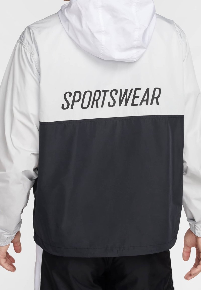 Czarna i biała kurtka sportowa z kapturem, z napisem "SPORTSWEAR" na plecach. Wykonana z lekkiego, gładkiego materiału.